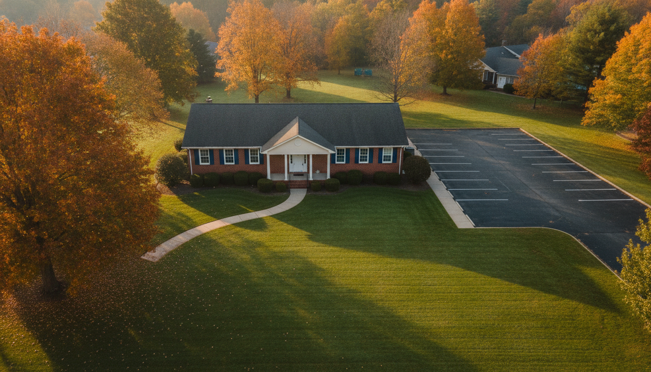 Aerial view of a single-story brick office with navy shutters, an empty parking lot and autumn maple trees in early morning light